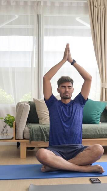 Uomo millenario positivo seduto su un tappetino e che pratica yoga in soggiorno