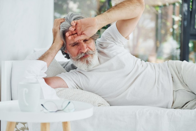 Uomo moderno ed elegante malato con capelli grigi e barba in casa sdraiato sul letto e si sente male