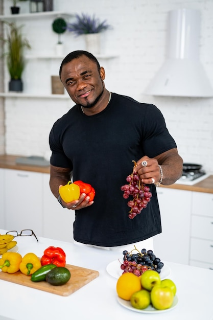 Uomo multirazziale in cucina pronto a preparare il pasto con frutta e verdura Il ragazzo tiene in mano uva e pepe Sfondo della cucina Cibo sano Vegani e vegetariani