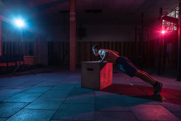 Uomo muscolare che allena le flessioni in scatola di legno in palestra scura Allenamento dell'atleta in luce al neon blu rossa Allenamento incrociato di uno stile di vita sano