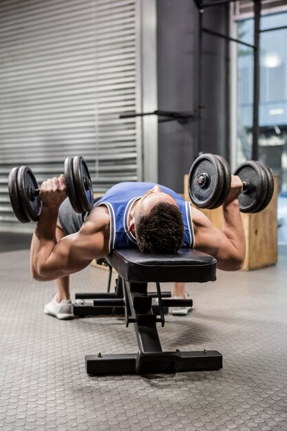 Uomo muscolare sulla panca sollevamento manubri presso la palestra di crossfit