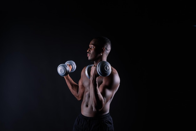 Uomo muscoloso africano isolato con manubri su sfondo scuro studio forte ragazzo nero senza camicia