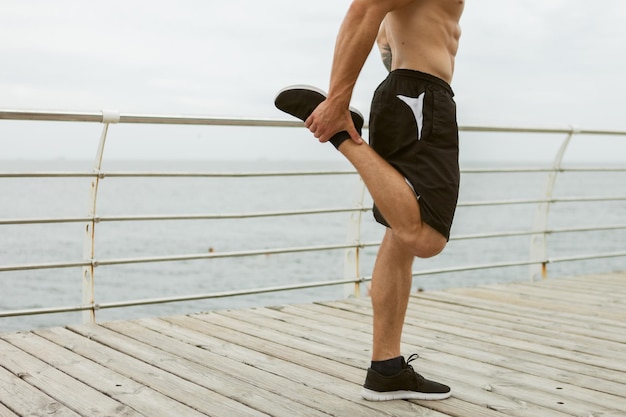 Uomo muscoloso con un torso nudo che fa allungamento delle gambe prima dell'allenamento sulla spiaggia
