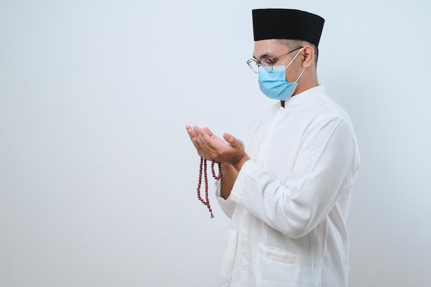 Uomo musulmano asiatico che indossa una maschera per il viso in preghiera a Dio al concetto di mese di digiuno di iftar timeramadan kareem sopra il muro bianco