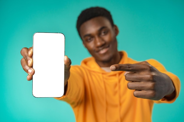 Uomo nero casual sorridente che punta allo schermo del telefono cellulare bianco vuoto in studio