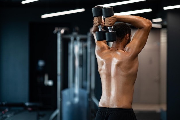 Uomo nero muscolare senza camicia che si allena con due manubri in palestra vista posteriore