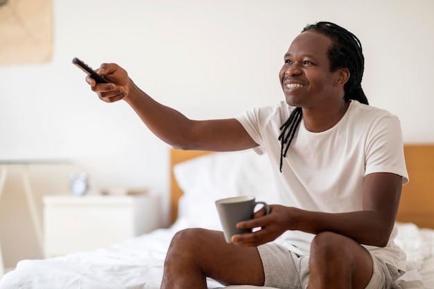 Uomo nero sorridente che beve caffè e guarda la TV mentre ci si rilassa a casa