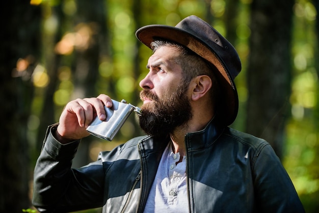 Uomo nomade portare alcol con sé Guy brutale cowboy barbuto bere alcol fiaschetta di metallo Hipster con barba bere alcol natura sfondo sfocato L'avventura inizia dal primo sorso Bevanda alcolica