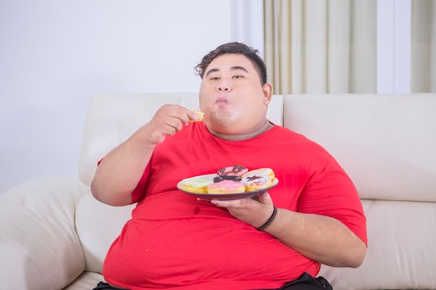 Uomo obeso che mangia un piatto di ciambelle sul divano