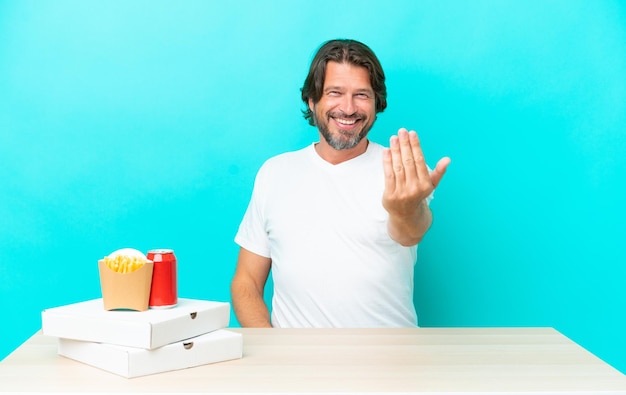 Uomo olandese anziano che mangia fast food seduto in un tavolo che invita a venire con la mano Felice che tu sia venuto