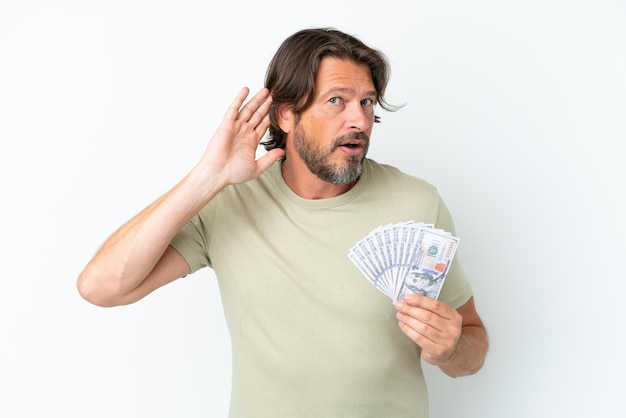 Uomo olandese anziano che prende un sacco di soldi isolato su sfondo bianco ascoltando qualcosa mettendo la mano sull'orecchio