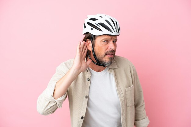 Uomo olandese anziano con casco da bici isolato su sfondo rosa ascoltando qualcosa mettendo la mano sull'orecchio