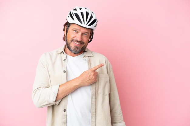 Uomo olandese anziano con casco da bici isolato su sfondo rosa che punta di lato per presentare un prodotto