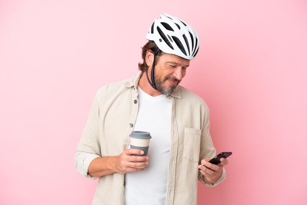 Uomo olandese anziano con casco da bici isolato su sfondo rosa che tiene caffè da portare via e un cellulare