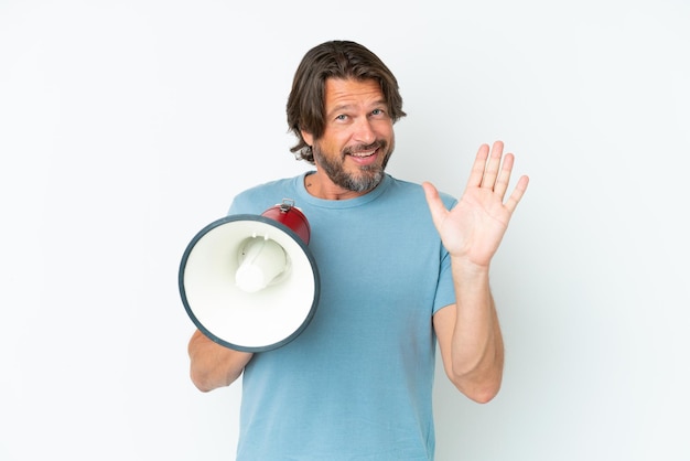 Uomo olandese anziano isolato su sfondo bianco che tiene in mano un megafono e saluta con la mano con espressione felice