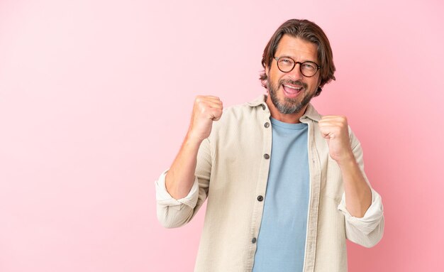 Uomo olandese anziano isolato su sfondo rosa che celebra una vittoria