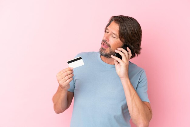 Uomo olandese anziano isolato su sfondo rosa che mantiene una conversazione con il telefono cellulare e tiene una carta di credito