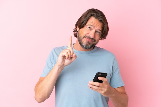 Uomo olandese anziano isolato su sfondo rosa utilizzando il telefono cellulare e sollevando il dito