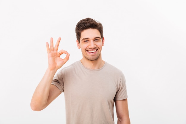 Uomo ottimista che ha setola e capelli castani che mostrano segno giusto sulla macchina fotografica con lo sguardo sincero, isolato sopra la parete bianca