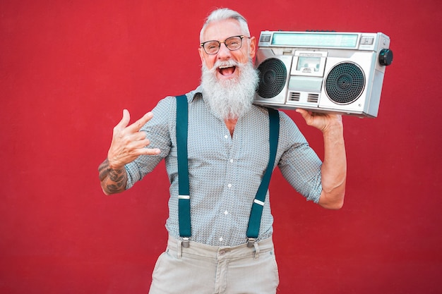 Uomo pazzo senior con stereo boombox anni '80 che suona musica rock con sfondo rosso - Ragazzo maturo alla moda divertendosi ballando con la radio vintage - Concetto di stile di vita allegro gioioso - Focus sul suo volto