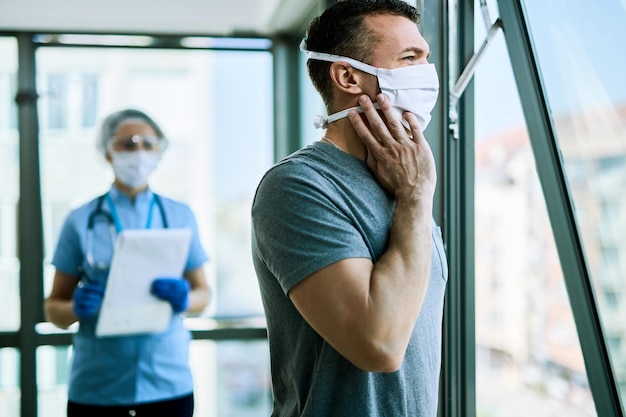 Uomo pensieroso con maschera facciale che sente i sintomi del COVID19 e guarda attraverso la finestra mentre si trova in clinica medica L'infermiera è in piedi sullo sfondo