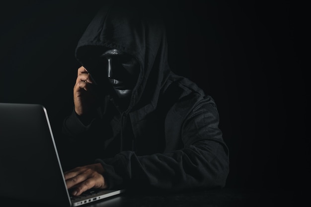 Uomo pericoloso hacker anonimo in incappucciato e maschera utilizzando computer e smartphone sul nero