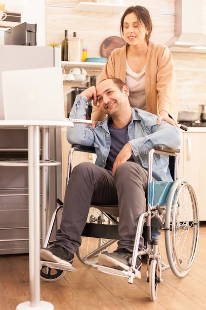 Uomo positivo in sedia a rotelle e moglie che guardano il laptop in cucina. Disabile paralizzato uomo handicappato con disabilità motoria che si integra dopo un incidente.