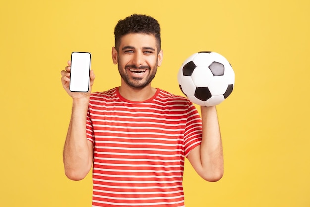 Uomo positivo sorridente con la barba in maglietta a righe che tiene e mostra smartphone con display bianco e pallone da calcio, scommettendo sul calcio. Studio indoor girato isolato su sfondo giallo