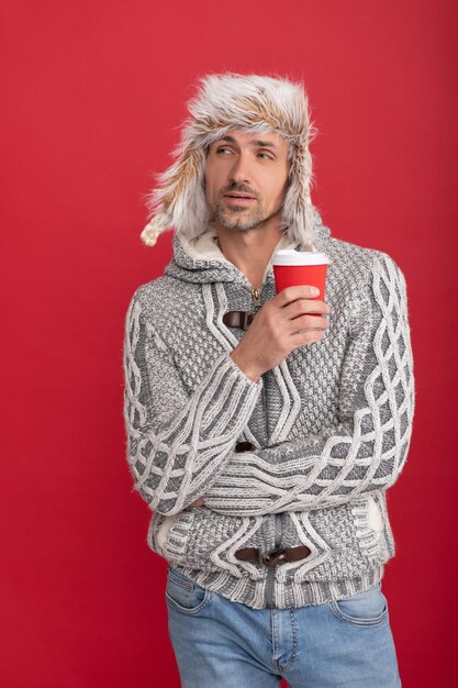 Uomo premuroso in maglione e paraorecchie che beve caffè dalla tazza su sfondo rosso, stile di vita
