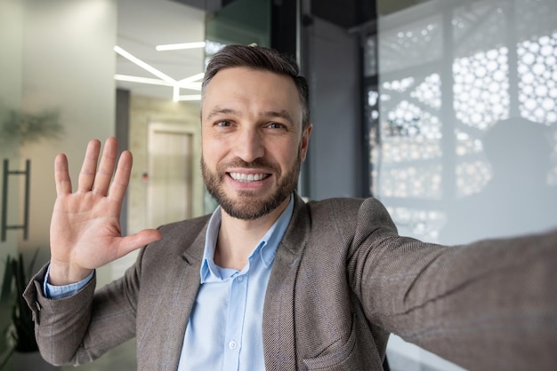 Uomo professionista allegro in abito che saluta qualcuno tramite una videochiamata che saluta mentre tiene la sua