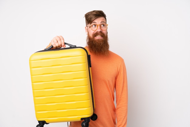 Uomo rossa con la barba lunga sul muro bianco in vacanza con la valigia di viaggio