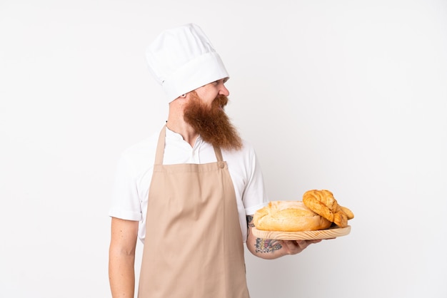 Uomo rossa in uniforme da chef. Panettiere maschio che tiene una tavola con parecchi pani con l'espressione felice