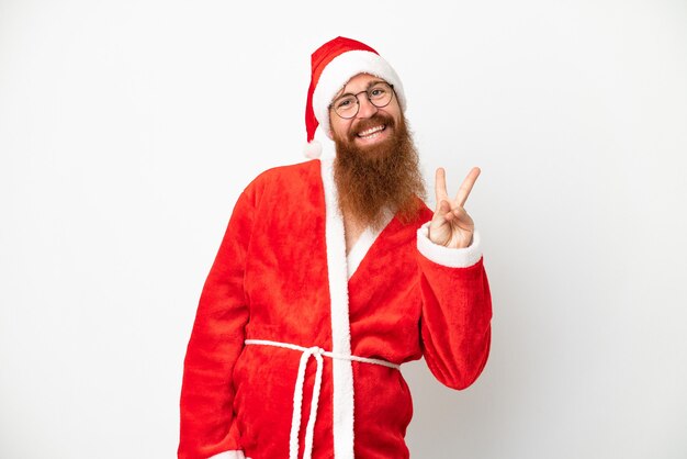 Uomo rossastro travestito da Babbo Natale isolato su bianco sorridente e che mostra il segno della vittoria