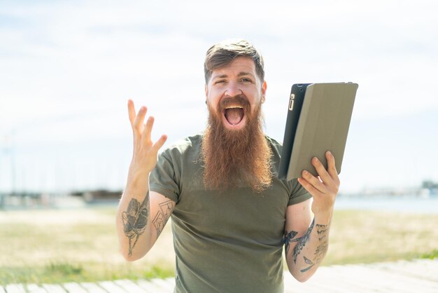 Uomo rosso con barba che tiene un tablet all'aperto con espressione facciale scioccata