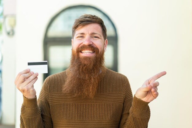 Uomo rosso con barba in possesso di una carta di credito all'aperto sorpreso e puntando il dito di lato