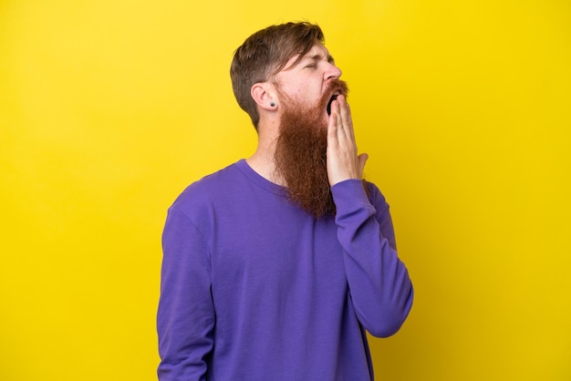 Uomo rosso con barba isolato su sfondo giallo che sbadiglia e copre la bocca spalancata con la mano