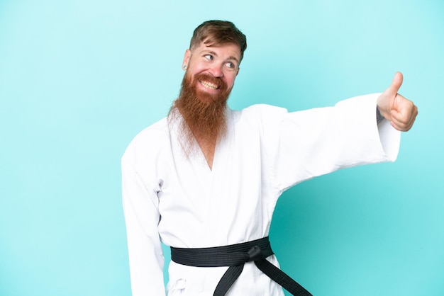 Uomo rosso con barba lunga che fa karate isolato su sfondo blu che dà un gesto del pollice in alto