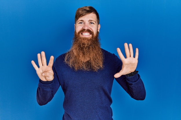 Uomo rosso con barba lunga che indossa un maglione blu casual su sfondo blu che mostra e punta verso l'alto con le dita numero nove mentre sorride fiducioso e felice.