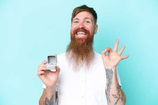 Uomo rosso con barba lunga che tiene un anello di fidanzamento isolato su sfondo blu che mostra il segno ok con le dita