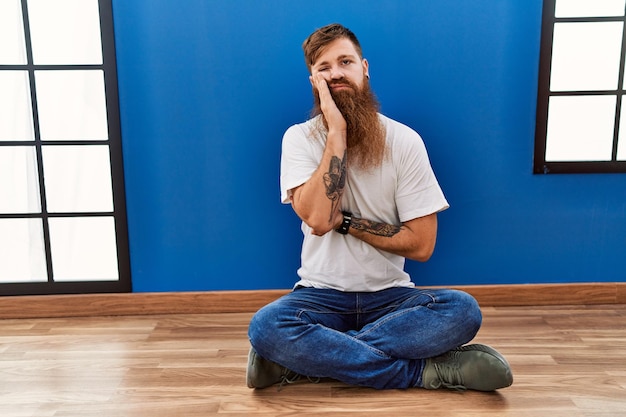 Uomo rosso con barba lunga seduto sul pavimento in una stanza vuota pensando che sembra stanco e annoiato da problemi di depressione con le braccia incrociate.