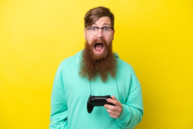 Uomo rosso con la barba che gioca con un controller per videogiochi isolato su sfondo giallo con espressione facciale sorpresa e scioccata