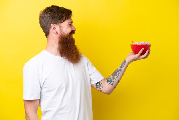 Uomo rosso con la barba che mangia una ciotola di cereali isolata su sfondo giallo con espressione felice