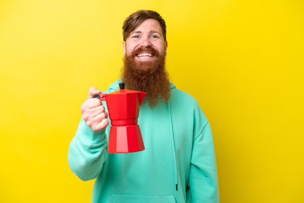 Uomo rosso con la barba che tiene la caffettiera isolata su sfondo giallo con espressione felice