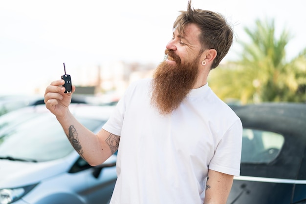 Uomo rosso con la barba che tiene le chiavi della macchina all'aperto con felice espressione