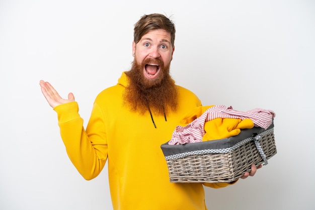 Uomo rosso con la barba che tiene un cesto di vestiti isolato su sfondo bianco con espressione facciale scioccata