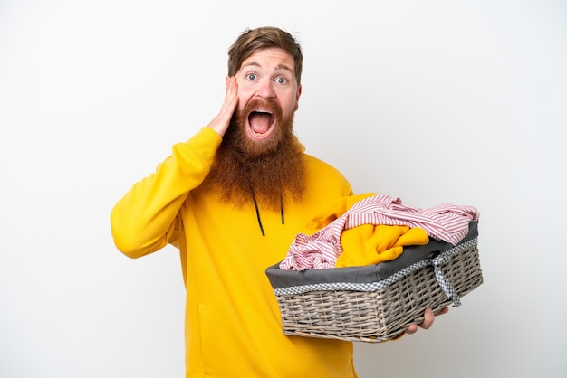 Uomo rosso con la barba che tiene un cesto di vestiti isolato su sfondo bianco con espressione facciale sorpresa e scioccata