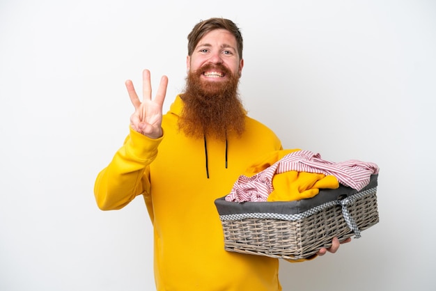 Uomo rosso con la barba che tiene un cesto di vestiti isolato su sfondo bianco felice e contando tre con le dita