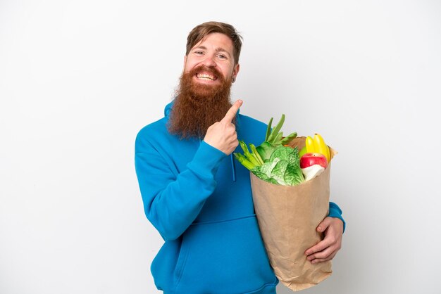 Uomo rosso con la barba che tiene un sacchetto della spesa isolato su sfondo bianco che indica il lato per presentare un prodotto