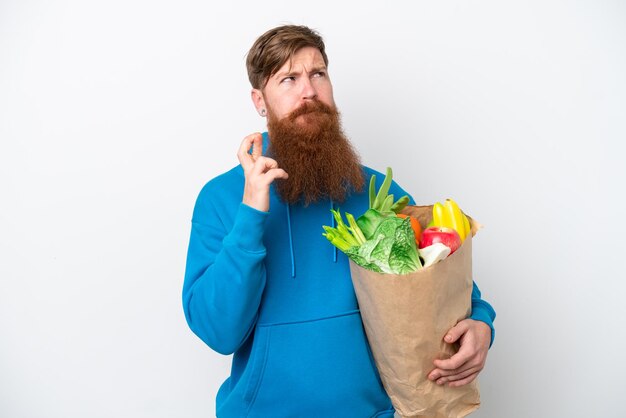 Uomo rosso con la barba che tiene un sacchetto della spesa isolato su sfondo bianco con le dita incrociate e augurando il meglio