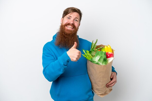 Uomo rosso con la barba che tiene una borsa della spesa isolata su sfondo bianco che dà un gesto del pollice in alto